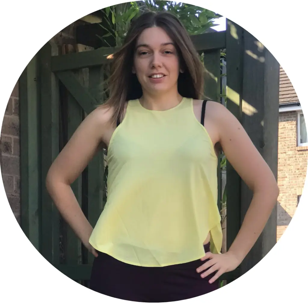 health coach UK: Woman stood outside with hands on hips. Wearing yellow tank top and dark maroon leggings.