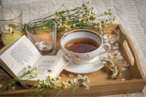 wooden tray with a teacup and saucer filled with chamolile tea. The tray has an open book on and flower decorations.